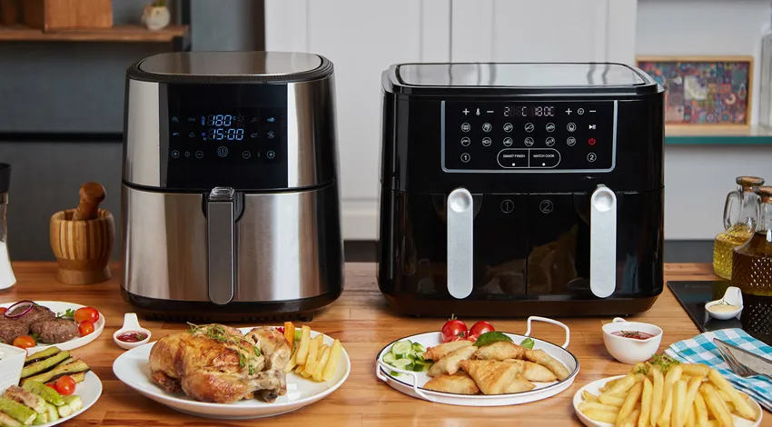 İki farklı airfryer pişirme makinesi yan yana ahşap bir masada yemeklerle birlikte duruyor.