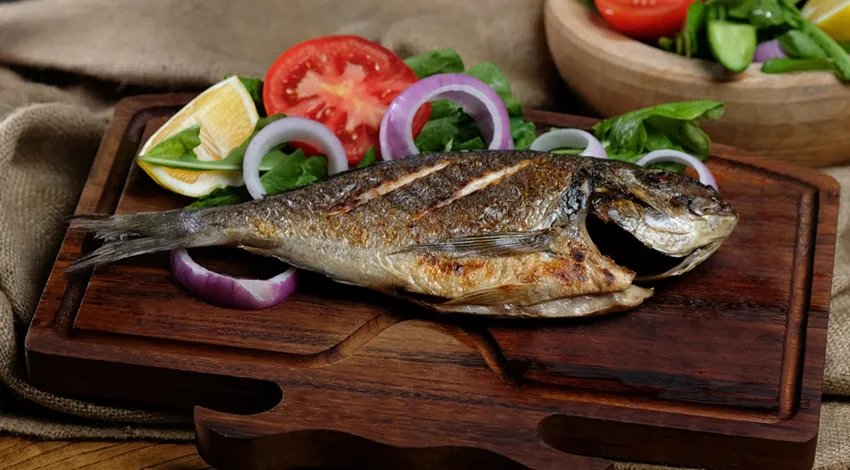 Ahşap sunum tahtası üzerinde ızgara balık, soğan halkaları, domates limon ve roka ile servis edilmiş