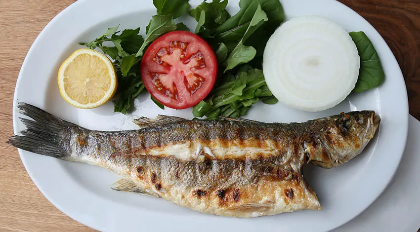 Izgara levrek beyaz tabakta yanında roka, soğan ve domates 