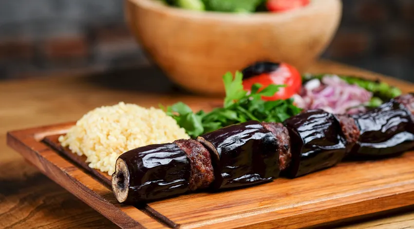 Patlıcanlı kebap, ahşap sunum tahtasında, bulgur pilavı, soğan, közlenmiş domates ve maydonoz ile servis edilmiş